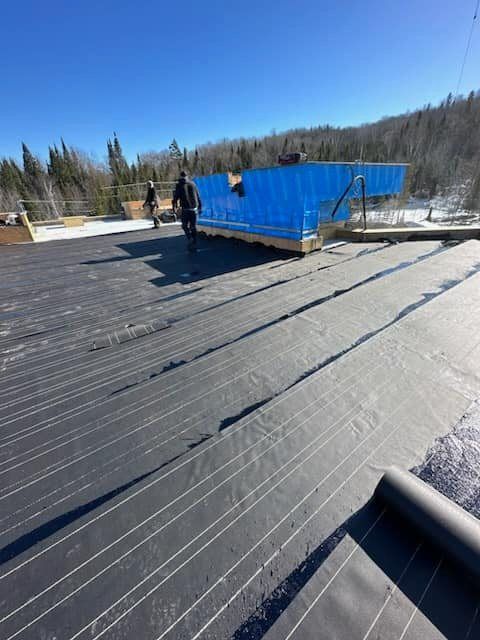 Deux personnes travaillent sur un toit plat en installant un revêtement de toiture noir à lignes blanches. L'isolant bleu est visible.