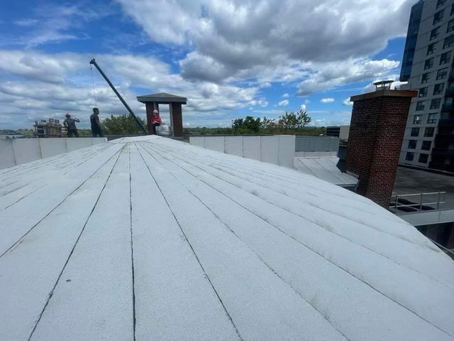 Des ouvriers sur un toit blanc sous un ciel nuageux, près d'une cheminée en briques et d'un bâtiment.
