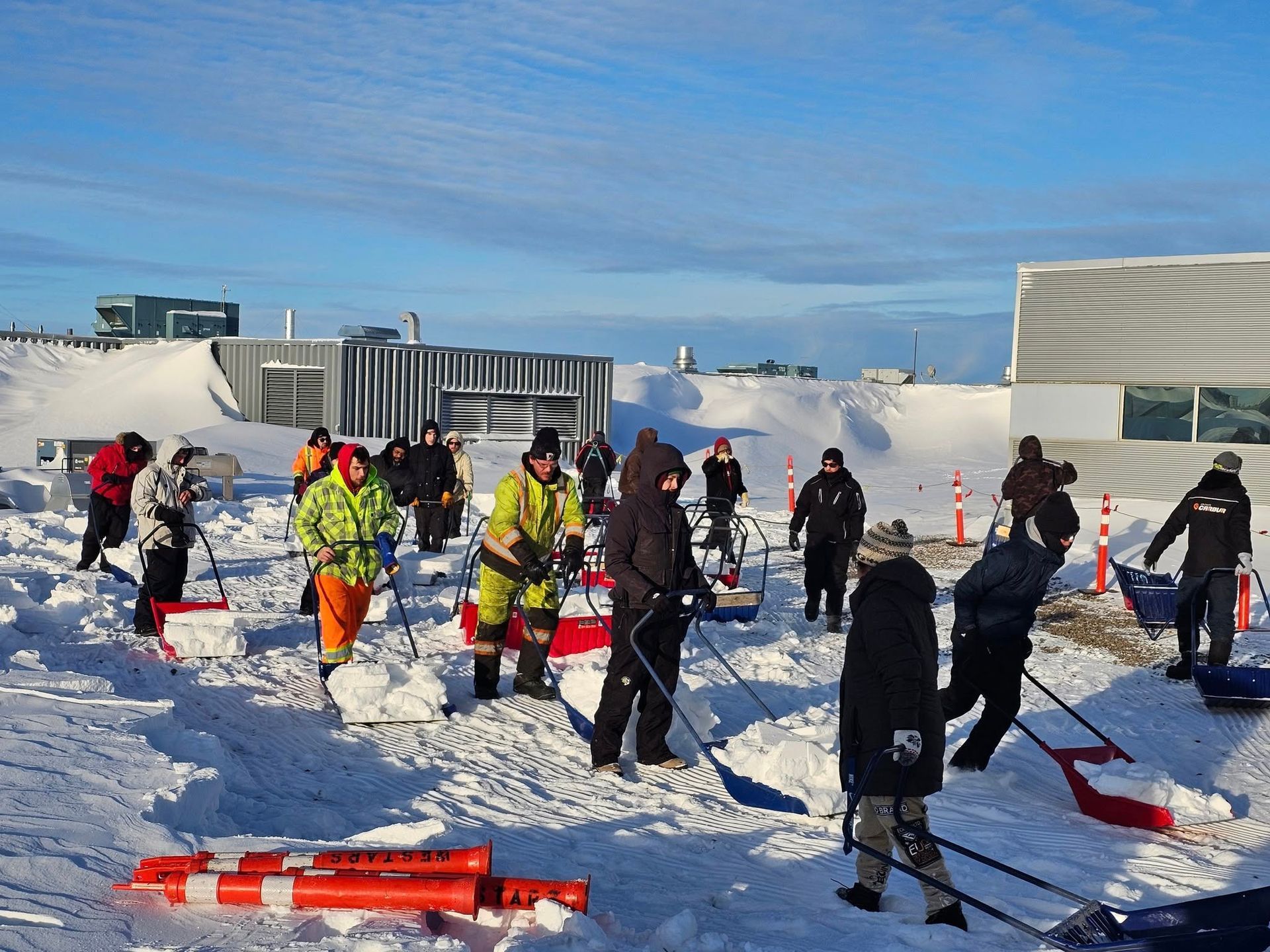 People shoveling snow outdoors on a bright, sunny day; white snow covers the ground and surrounding buildings.