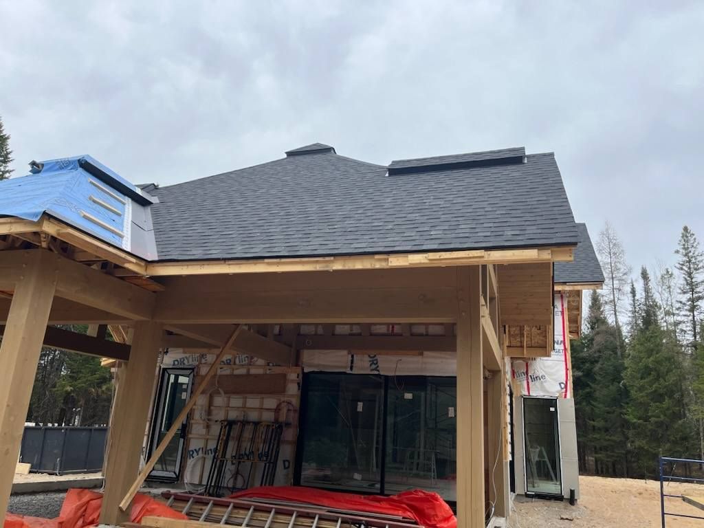 New house under construction with dark gray shingle roof, wooden frame, and wrap. Cloudy sky in background.