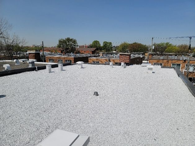 Toit plat recouvert de gravier blanc, avec des bâtiments en briques et des arbres en arrière-plan sous un ciel bleu clair.