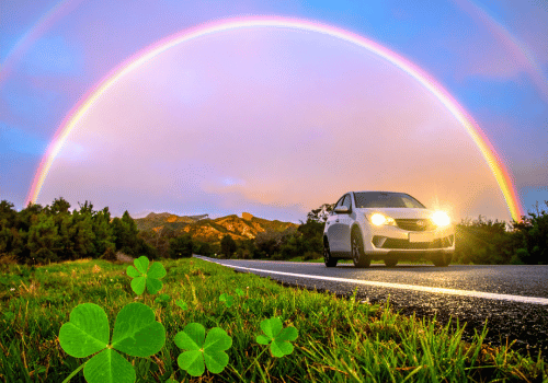 Car driving on a scenic road beneath a bright rainbow with shamrock graphics in the grass.