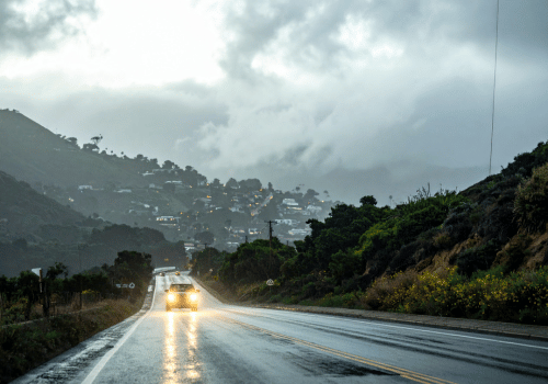 Car driving on a wet road in rainy hillside conditions with headlights reflecting on the pavement.