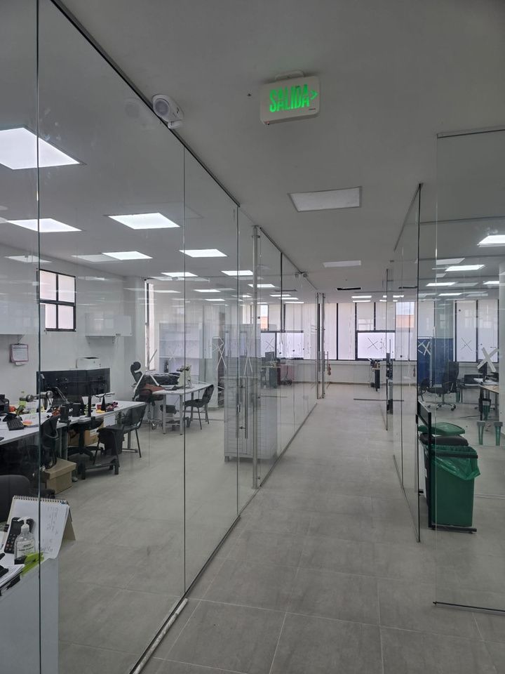 Interior de oficina con salas acristaladas, escritorios, sillas y trabajadores, iluminación brillante.