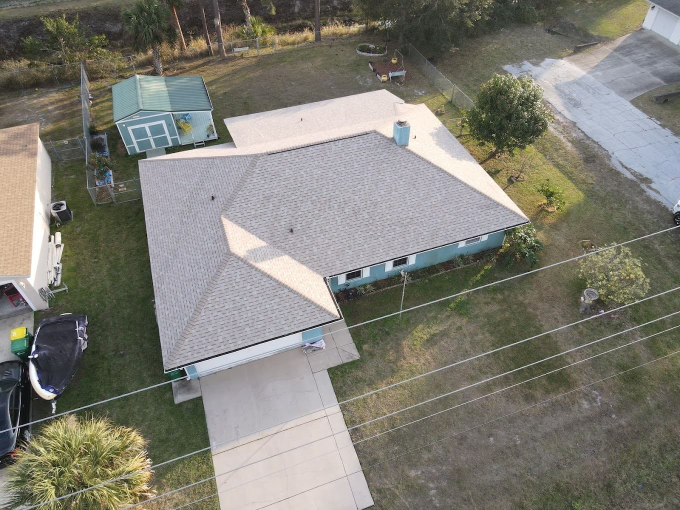 An aerial view of a single-story house with a light-colored roof, a concrete driveway, and a small green shed in the yard.