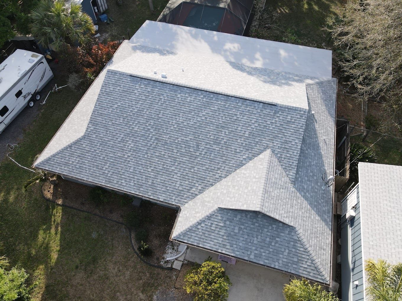 An aerial view shows a light-gray shingled roof on a residential home with a white trailer parked on the side.