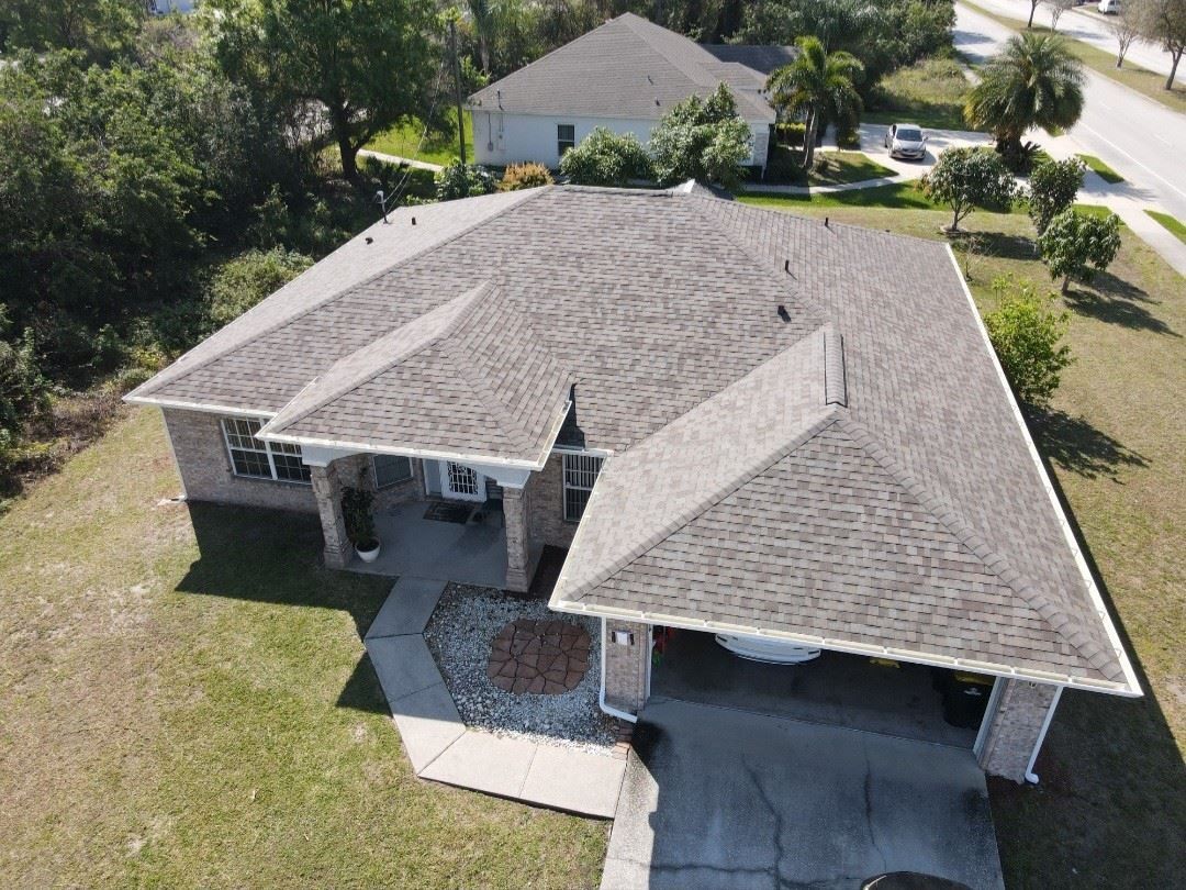 An aerial, high-angle view of a single-story house with a brown shingled roof, surrounded by trees and a paved driveway.