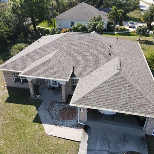 An aerial view of a suburban house with a gray shingled roof, a paved driveway, and a small front yard.