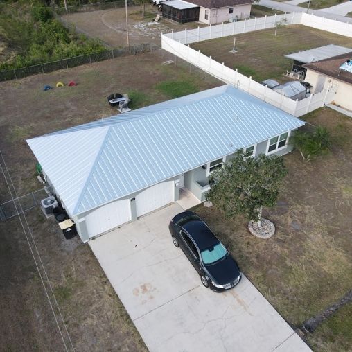 An aerial view of a single-story house with a light-colored metal roof and a driveway with a dark car parked in front.