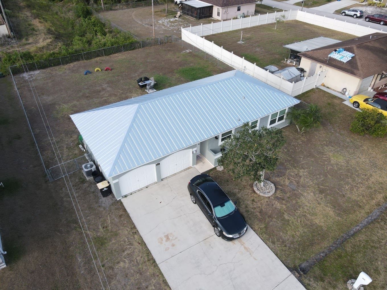 An aerial view of a single-story house with a light-colored metal roof and a driveway with a dark car parked in front.