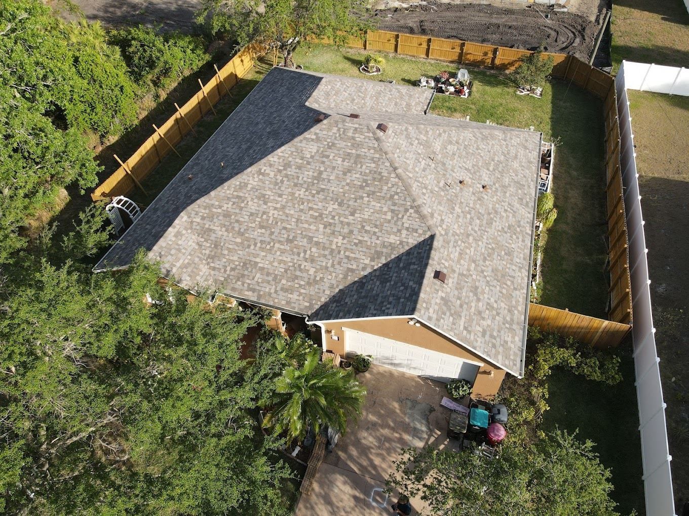 An aerial view of a suburban house with a gray shingled roof, a paved driveway, and a fenced-in backyard.