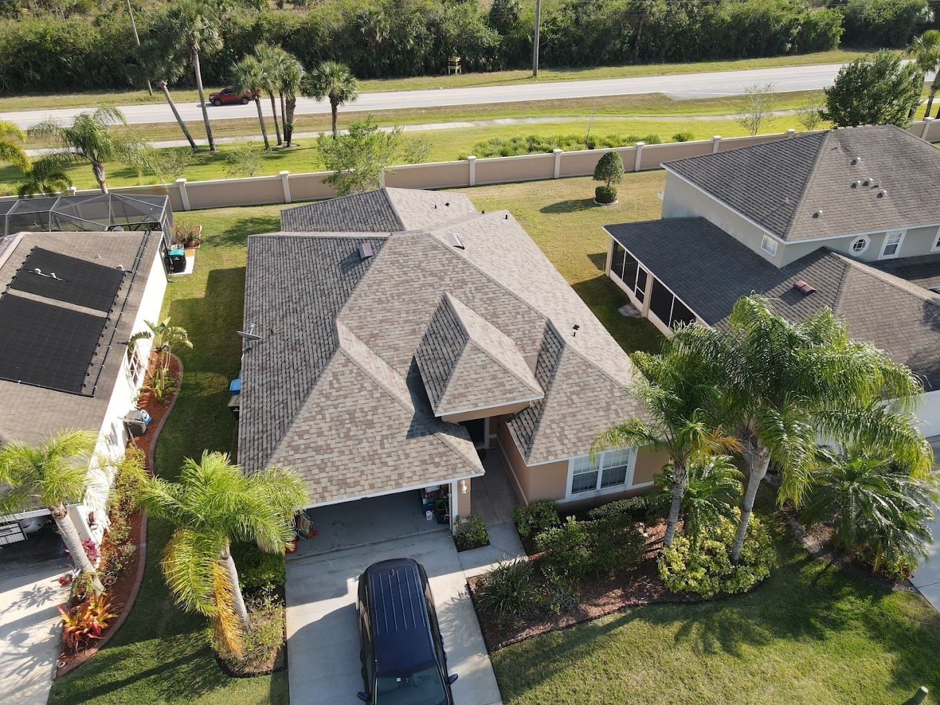 Aerial view of a residential suburban home with a gray shingled roof, driveway, and surrounding palm trees.