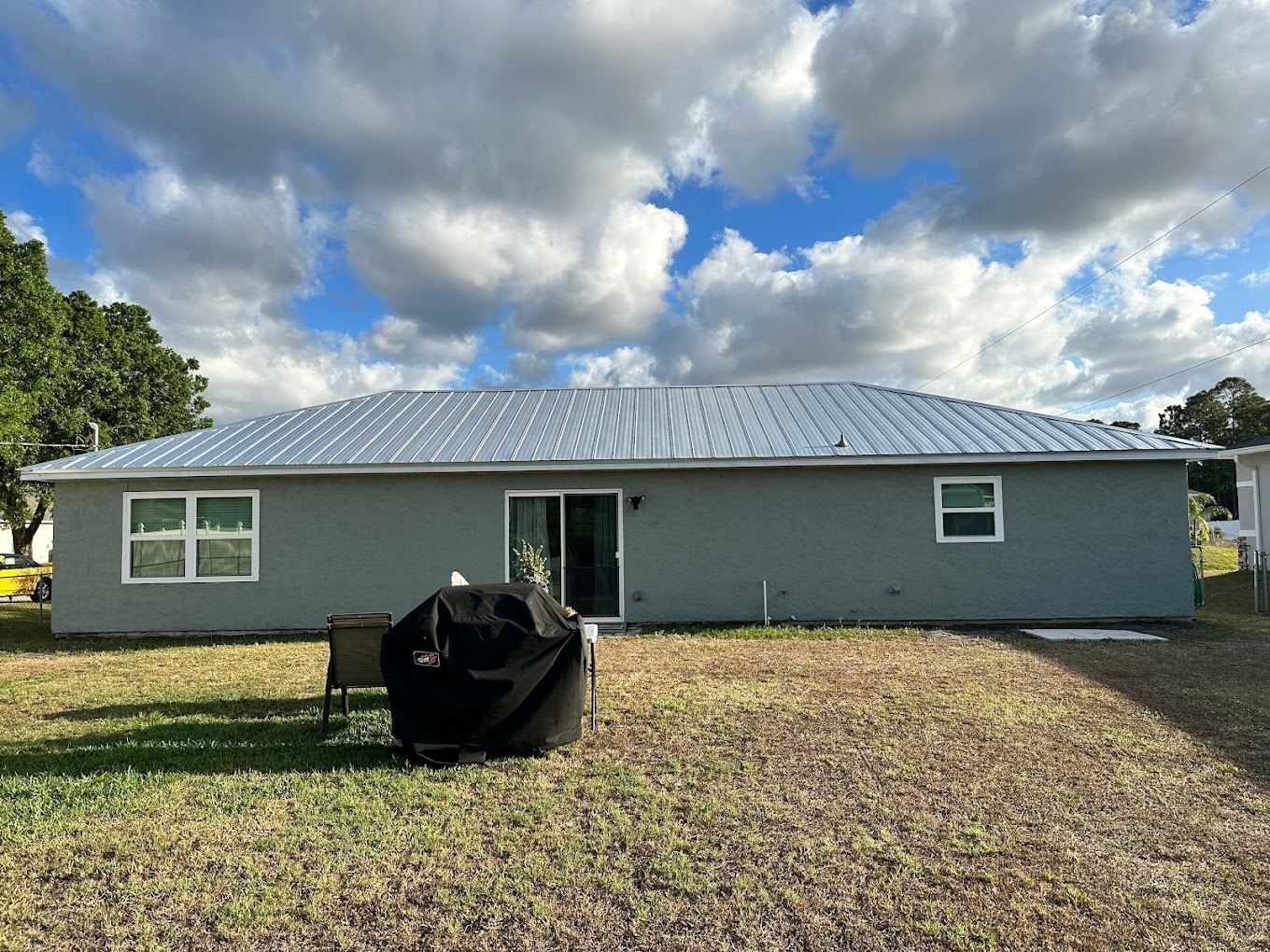 A gray single-story house with a metal roof and large backyard under a cloudy sky.