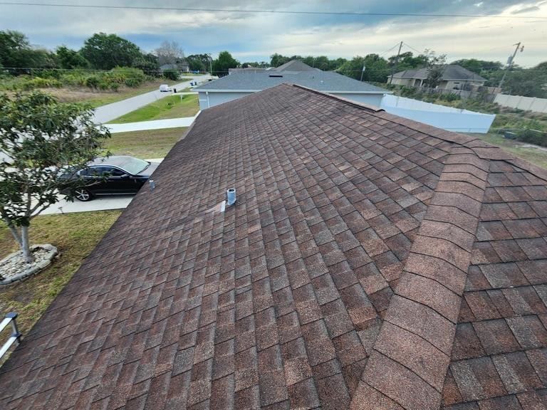 A view from a rooftop looking down onto brown asphalt shingles, a hip ridge, a metal vent, and a suburban street below.