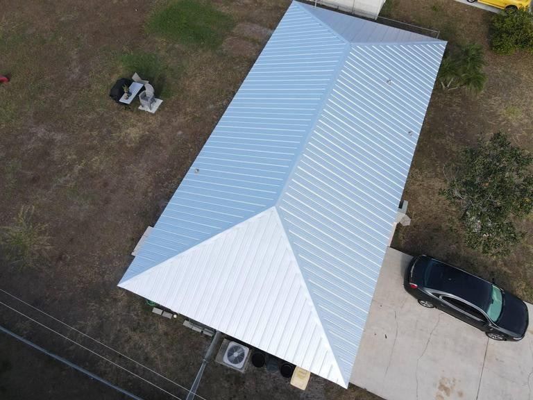 A high-angle aerial view of a rectangular house with a light grey metal hip roof, situated on a grassy lot with a car.