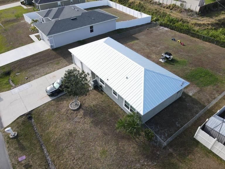 Aerial view of a light gray house with a white metal roof, a driveway with a car, and a grassy yard with a white fence.