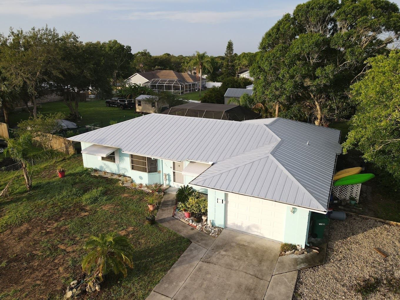 An aerial view of a pale blue house with a light gray metal roof, surrounded by trees and a concrete driveway.