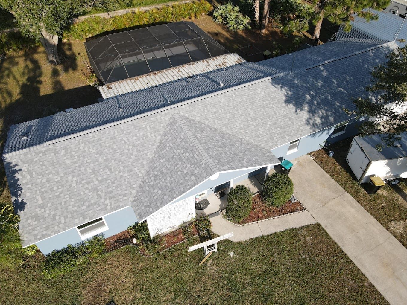 An aerial view of a single-story house with a light gray shingled roof, a dark enclosed patio, and a paved driveway.
