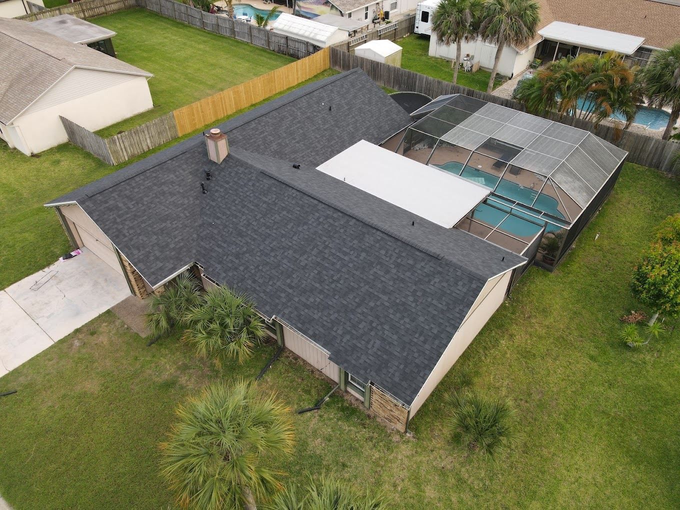 Aerial view of a residential house with a dark gray shingled roof, a white patio cover, and an enclosed screened-in pool.