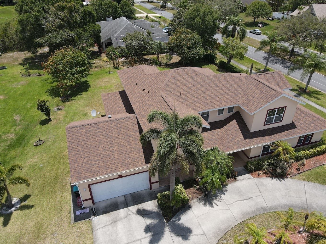 An aerial view of a tan two-story house with a brown shingled roof, a garage, and a driveway surrounded by trees and grass.