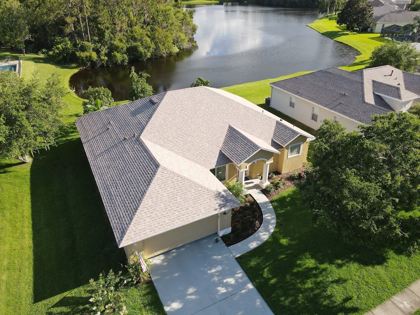 An aerial view of a suburban house with a gray shingled roof, a paved driveway, and a lake in the backyard.