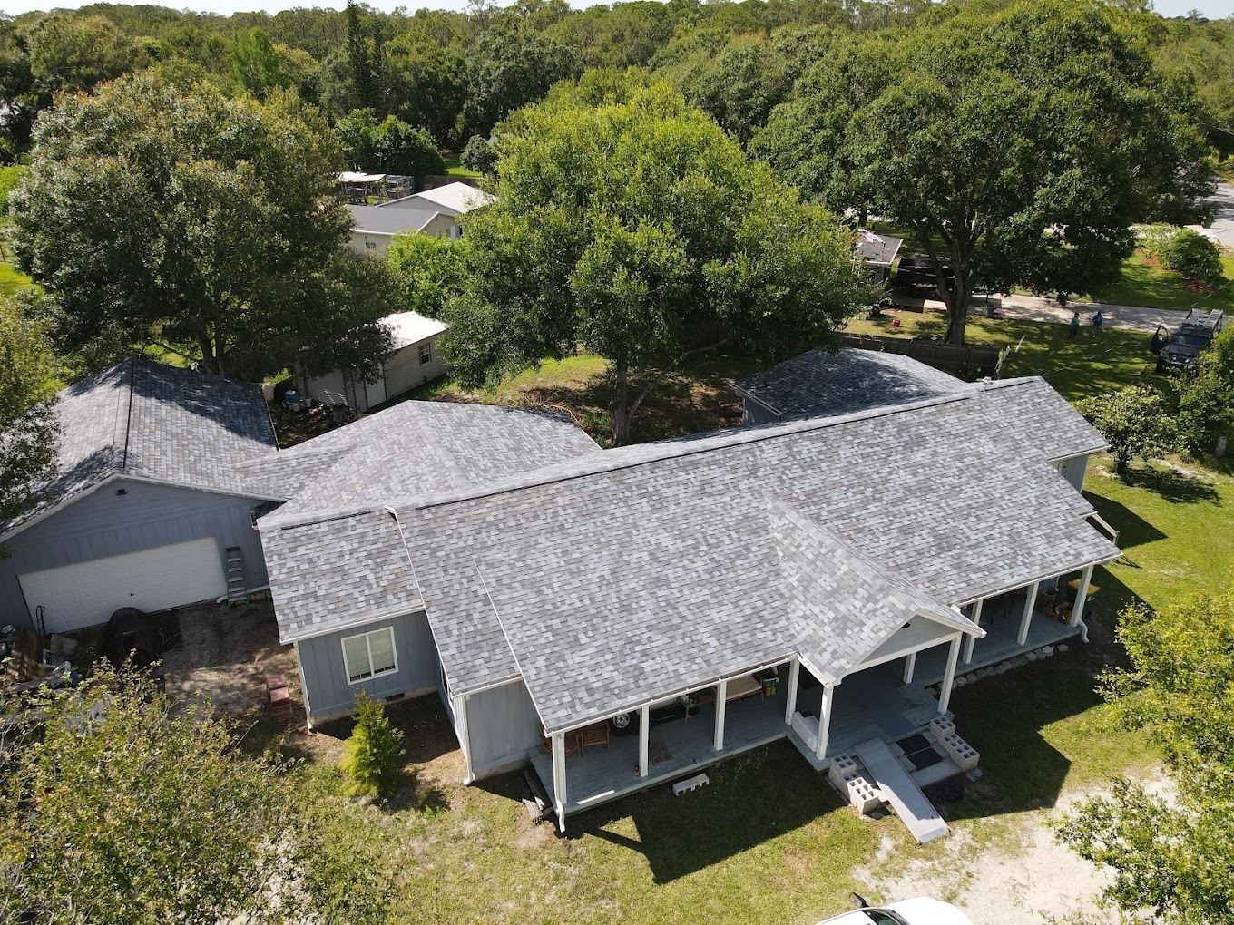 An aerial view of a single-story gray house with a shingled roof, a front porch, and an attached garage among trees.