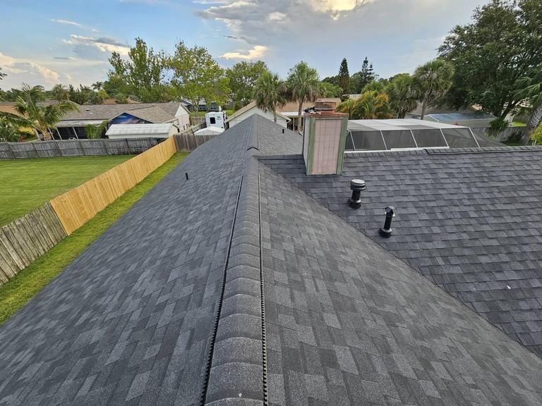 A high-angle view of a dark gray asphalt shingle roof, showing a ridge, a chimney, plumbing vents, and a suburban backyard.