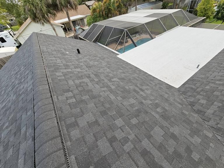 A view from a residential roof looking down at a gray shingled roof, a white flat roof section, and a screened pool cage.