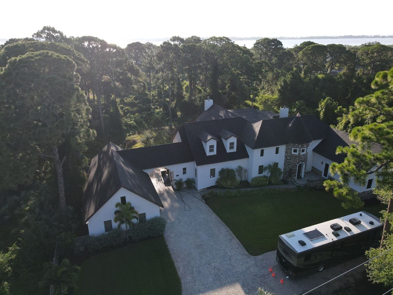 Aerial view of a white, multi-gabled house with a large gravel driveway, green lawn, and a parked motorhome near trees.