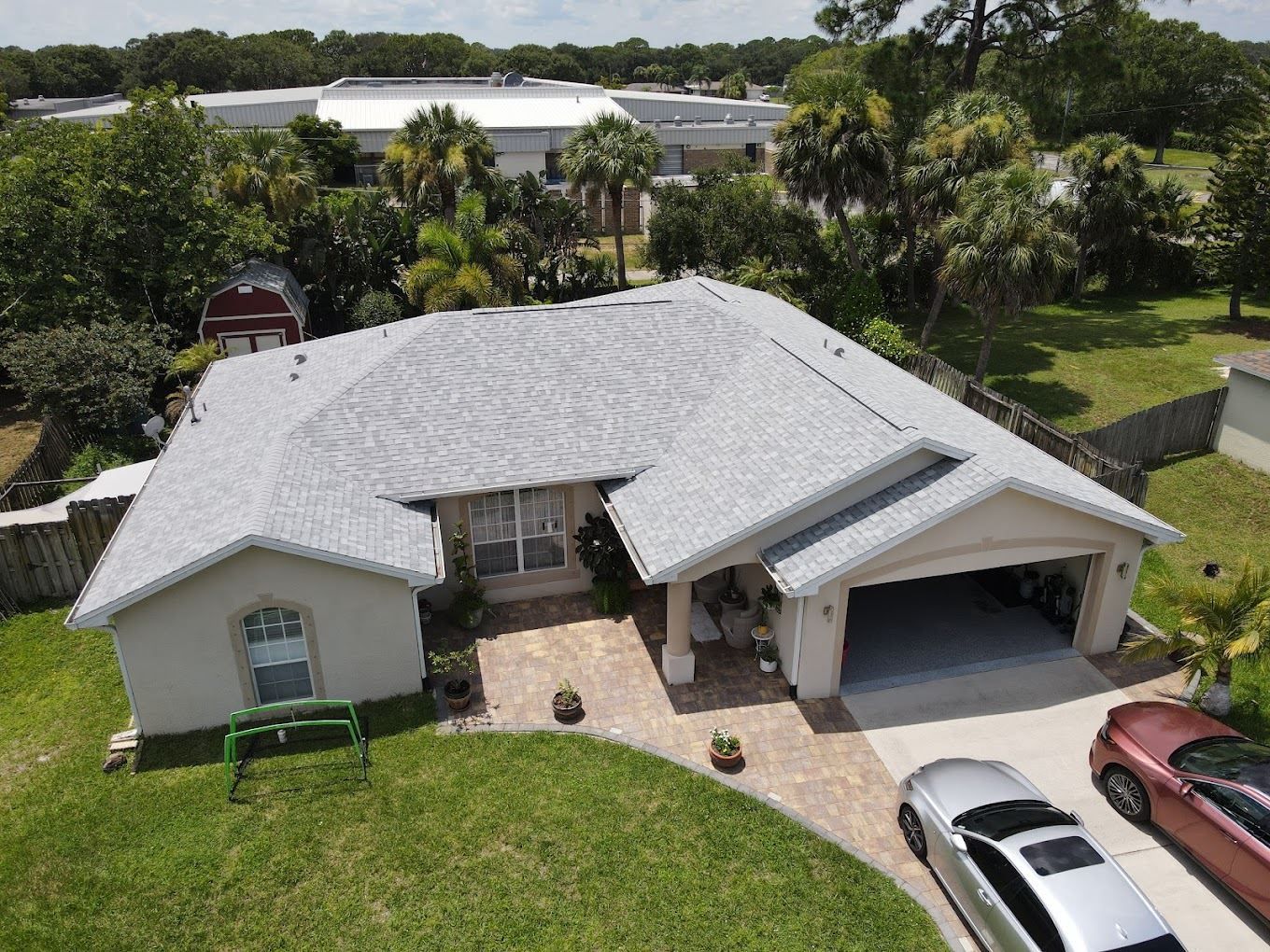 An aerial view of a single-story house with a light gray shingled roof, a paved driveway, and two parked cars.