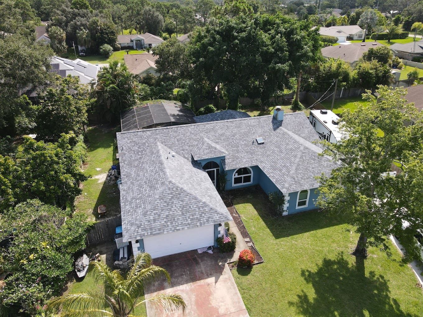 An aerial view of a blue, single-story ranch house with a gray shingled roof, surrounded by a lawn and mature trees.
