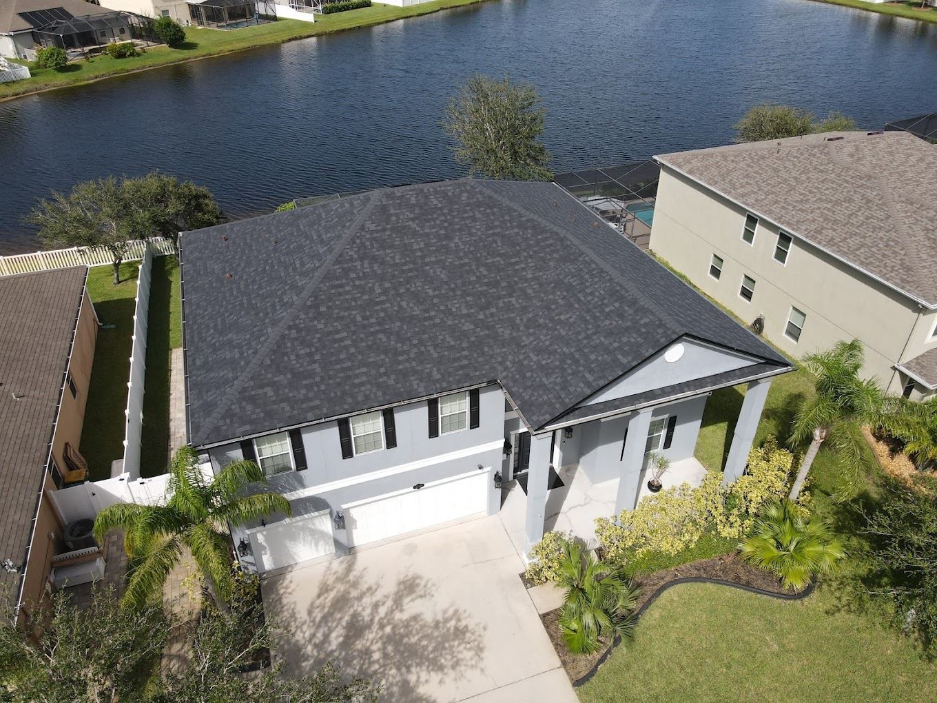 An aerial view of a gray suburban house with a dark roof and a two-car garage, situated next to a pond.