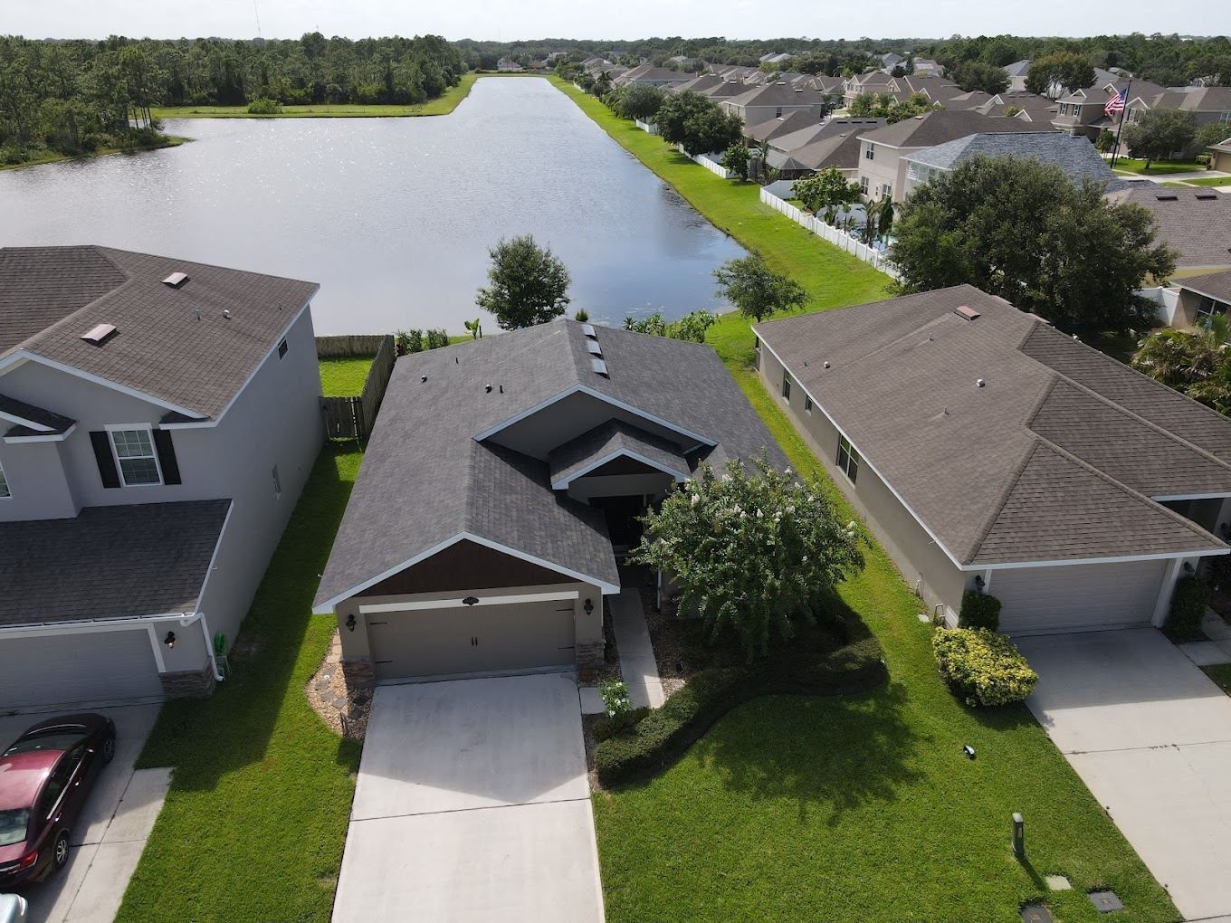An aerial view of a suburban neighborhood showing three houses with gray roofs situated alongside a long, narrow lake.
