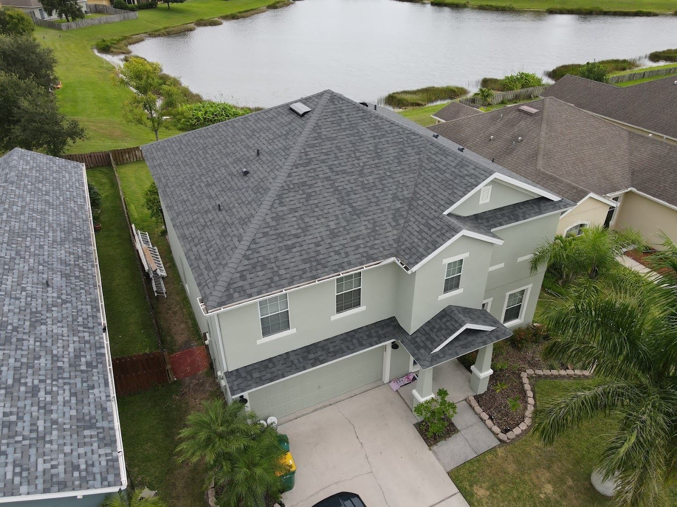 Aerial view of a light green two-story house with a gray shingled roof next to a lake and a neighboring house.