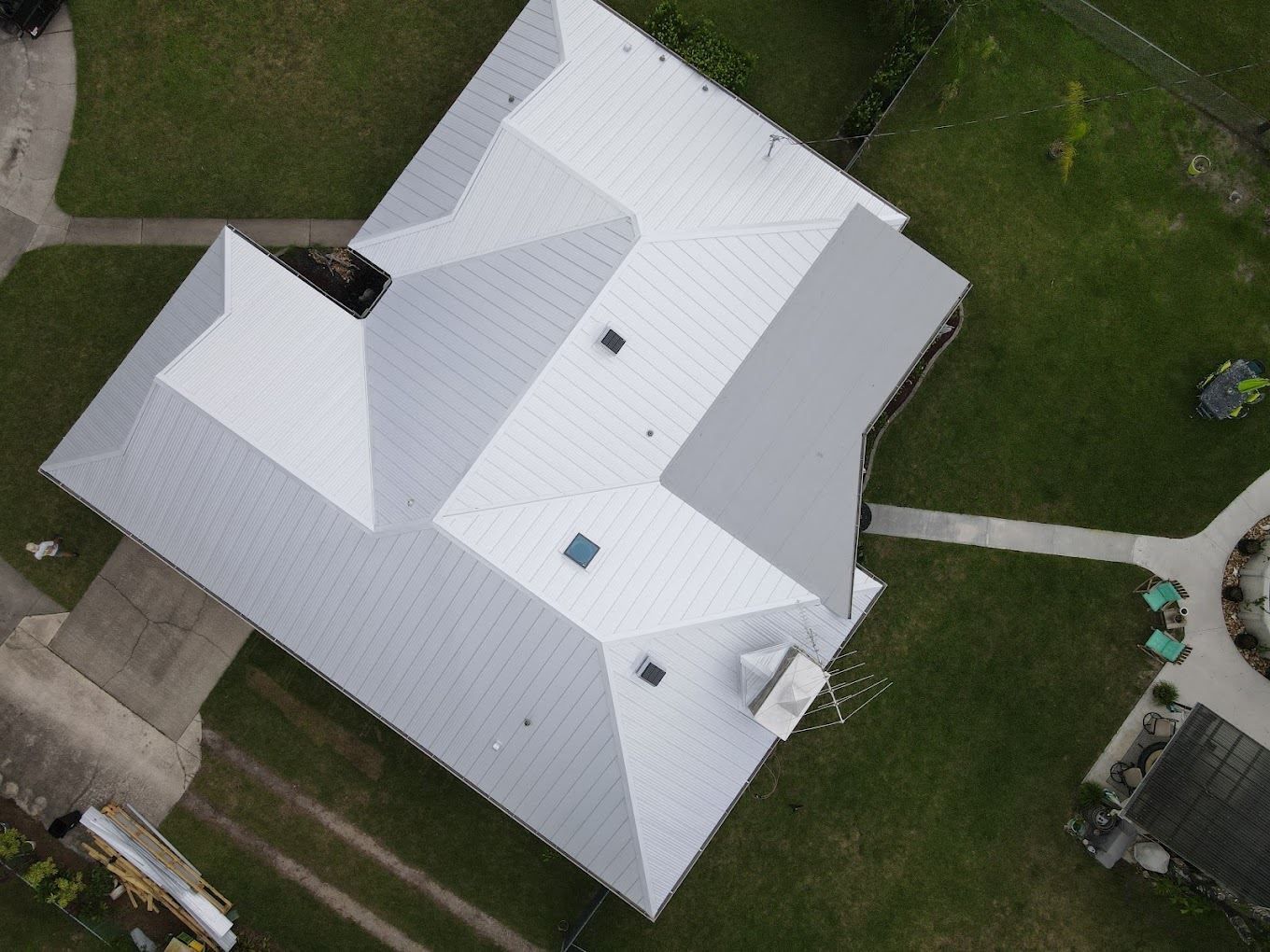 An aerial view of a house with a light gray metal roof, surrounded by green lawns and walkways.