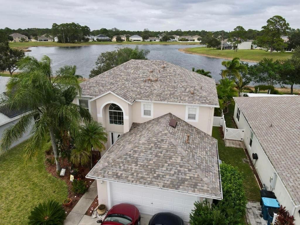An aerial view of a two-story beige house with a grey shingled roof, front driveway, and a lake in the background.