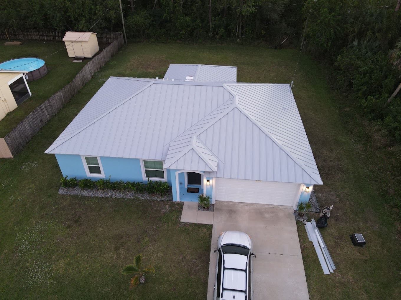 An aerial view of a light blue house with a gray metal roof, a driveway with a car, and a backyard pool and shed.