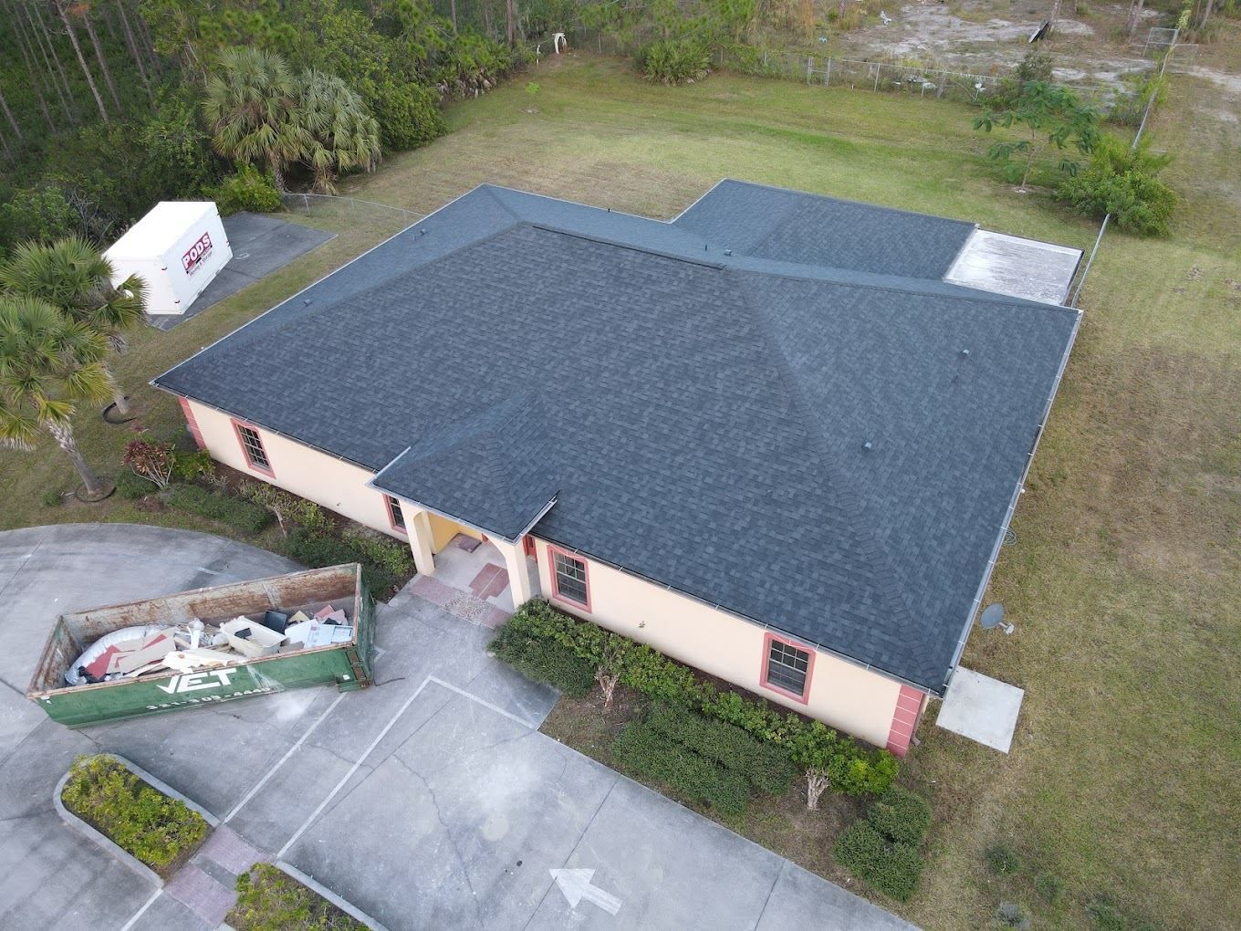 An aerial view of a tan, single-story building with a new dark gray shingled roof, next to a dumpster in a parking lot.