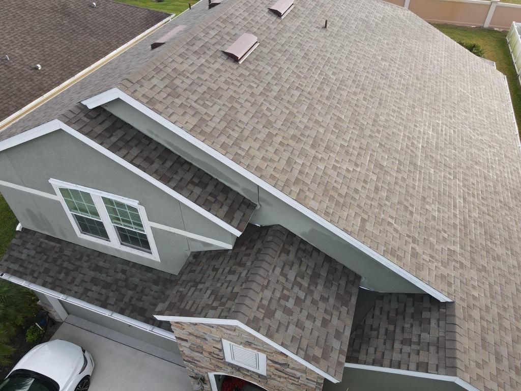 An elevated view of a house with a multi-textured, gray and tan shingled roof, featuring several peaks and small skylights.