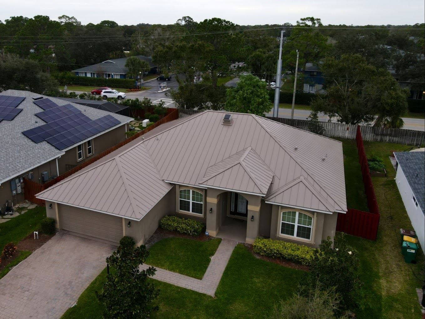 Aerial view of a tan, single-story suburban house with a metal roof and a paved driveway, surrounded by a lawn.