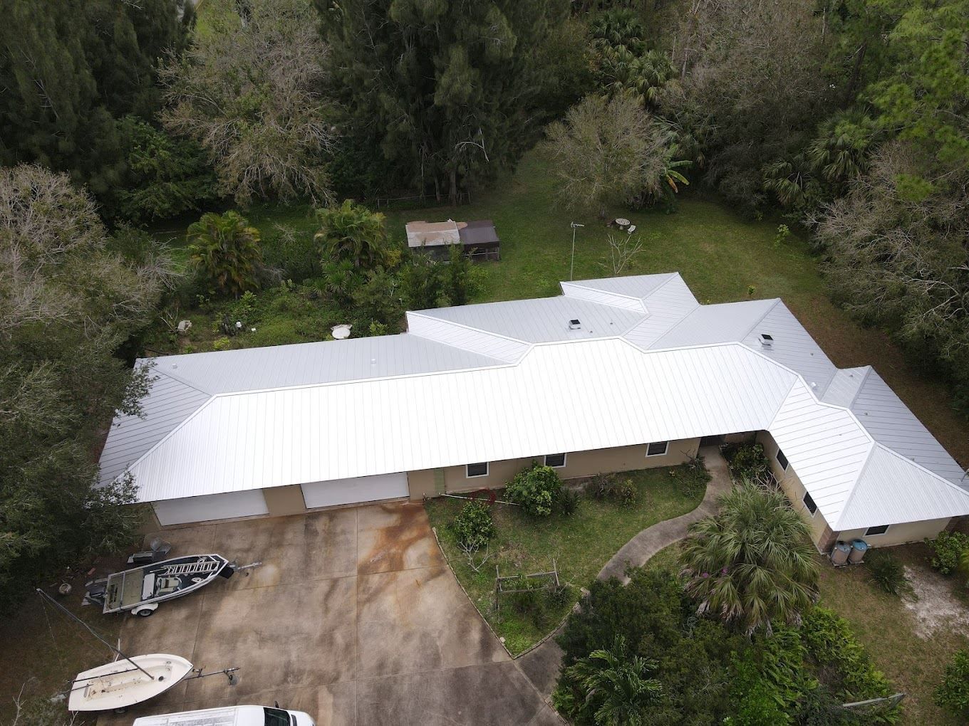 An aerial view of a suburban house with a white metal roof, a driveway with several boats, and surrounding trees.