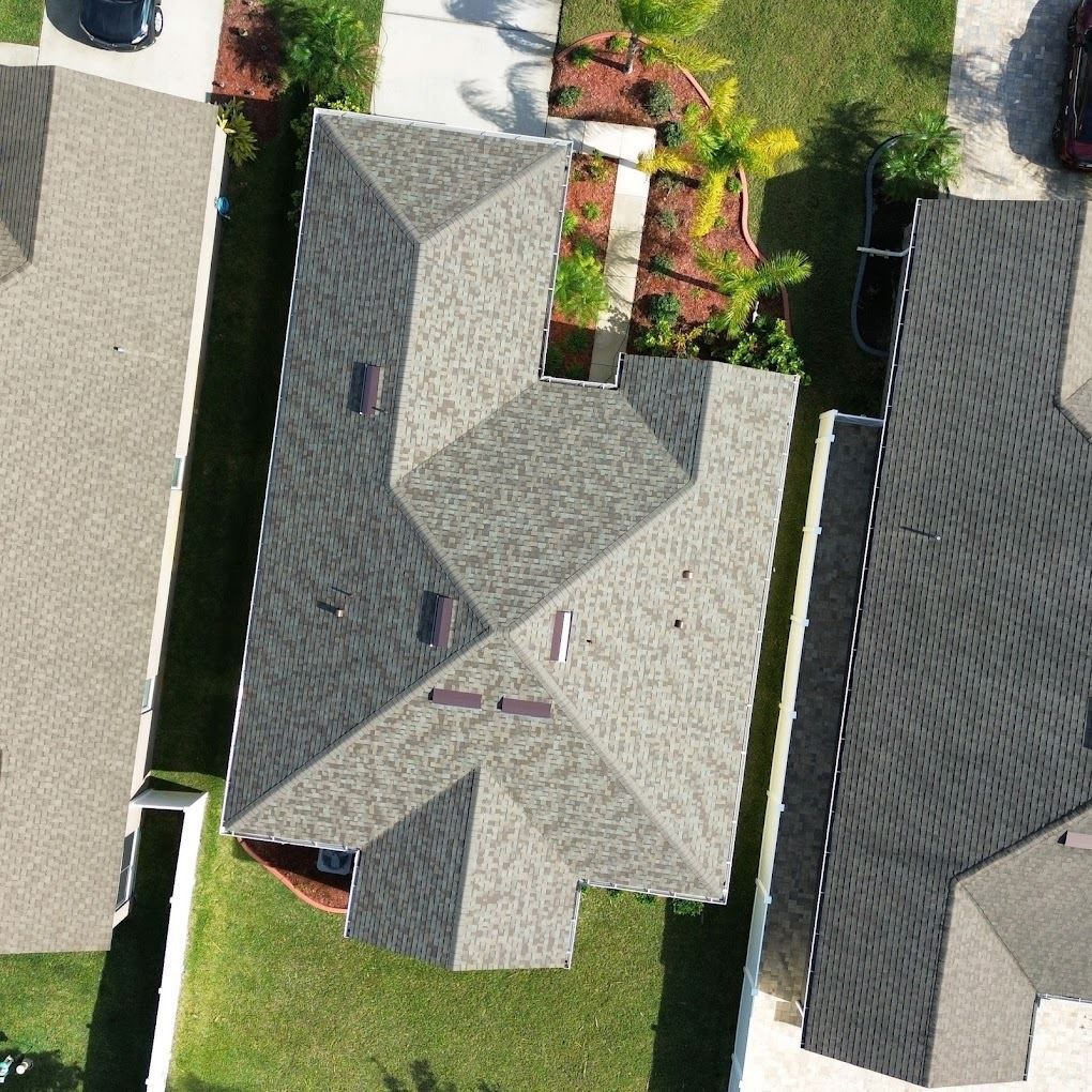 An aerial top-down view of a residential house with a complex shingled roof, surrounded by a lawn and adjacent properties.