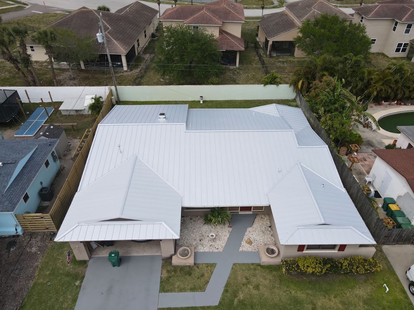 A high-angle view of a residential house with a light grey metal roof, a paved walkway, and a driveway in a suburban area.