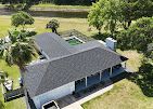 Aerial view of a gray-roofed residential house with a pool, patio, and surrounding trees near a pond.