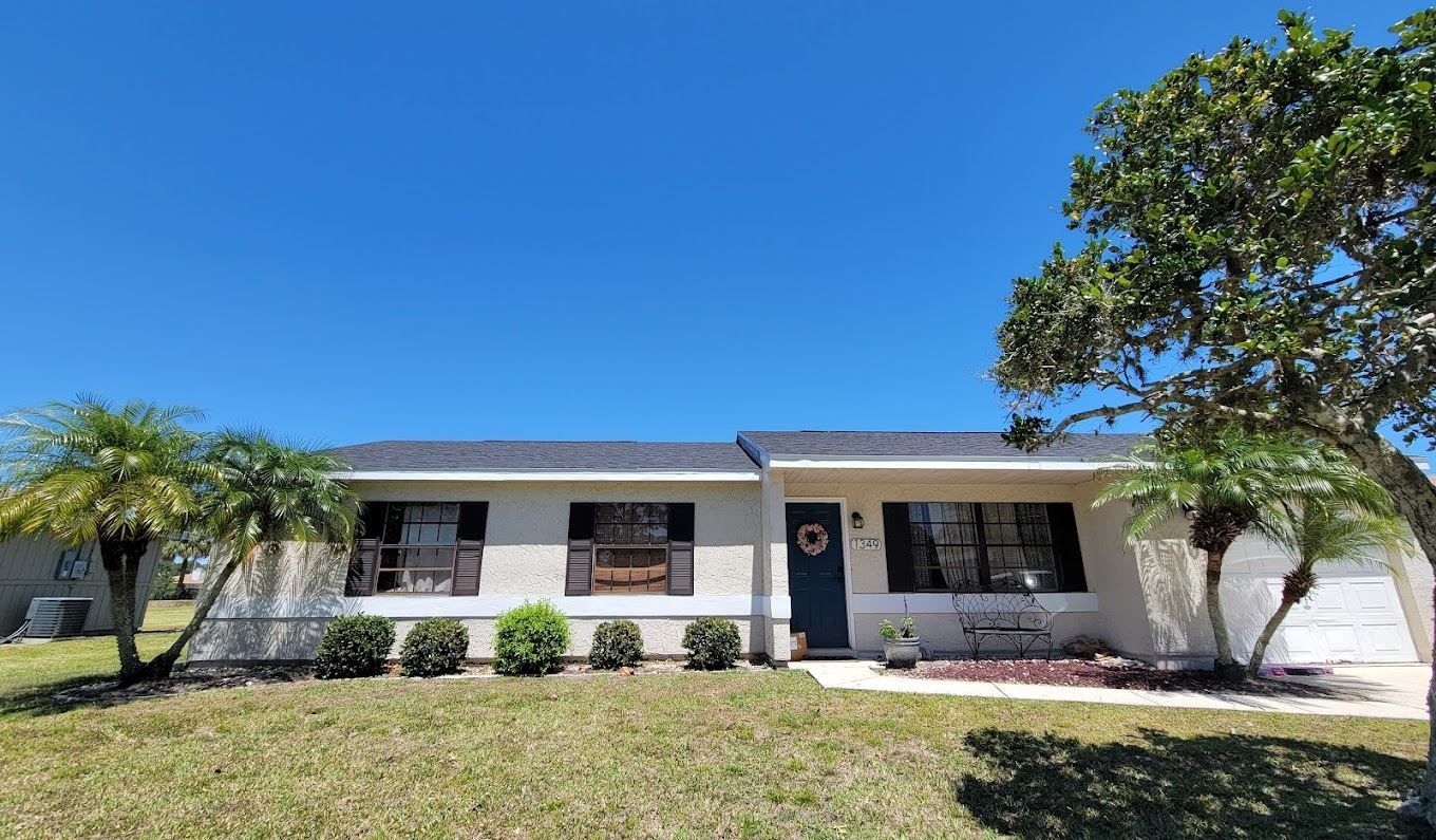 Single-story suburban house with a dark blue front door, light-colored walls, dark shutters, and palm trees in the yard.