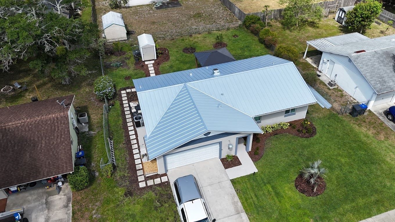 Aerial view of a residential house with a light blue roof, white garage, front walkway, and two small sheds in the yard.