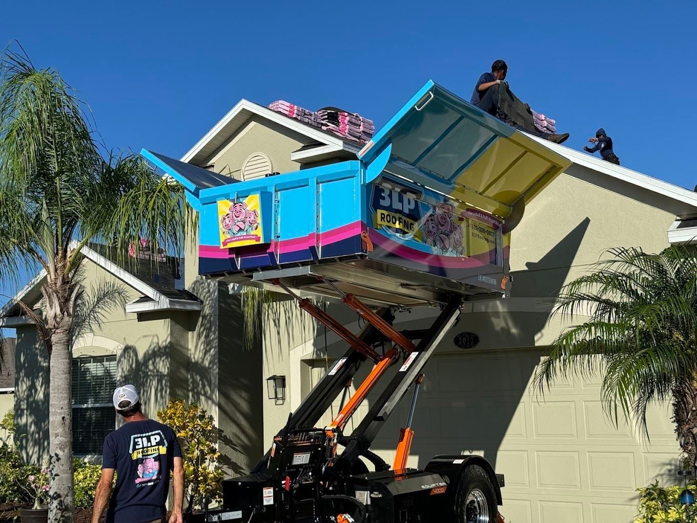 Workers use a bright blue hydraulic dump trailer raised against a house roof to collect old shingles for disposal.