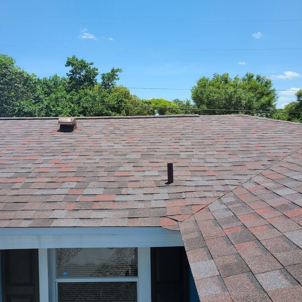 A high-angle view of a brown shingled roof under a clear blue sky, featuring a vent pipe and a chimney cap.
