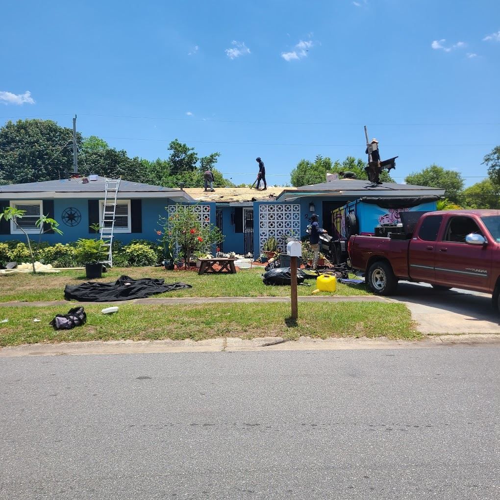 Workers replacing the roof of a blue house on a sunny day, with a red truck parked in the driveway.