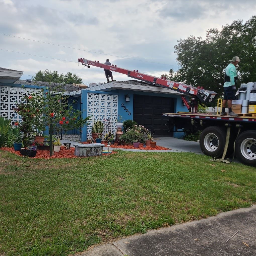 A worker uses a red roof-loading conveyor on a truck to deliver supplies to the roof of a blue house.